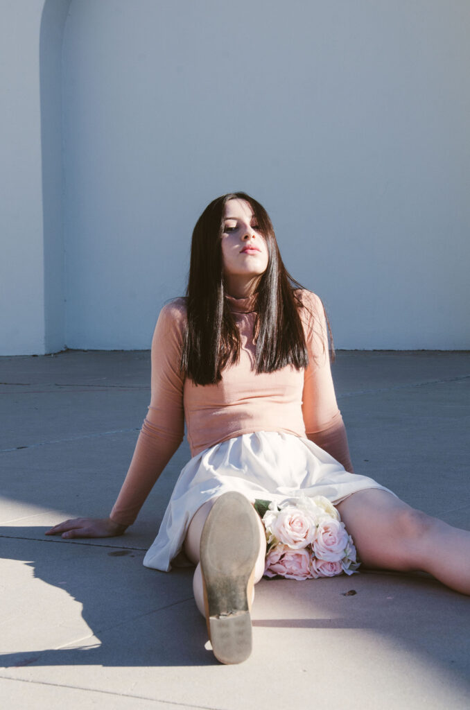 Gabriela Inzunza sits on the ground leaning back against a white wall with her eyes closed and a bouquet of flowers tucked into her skirt.